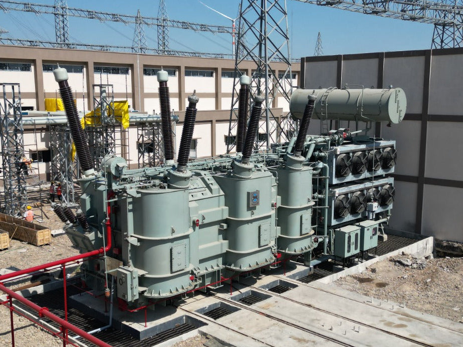 Large electrical transformer in an outdoor setting with power lines and structures in the background.