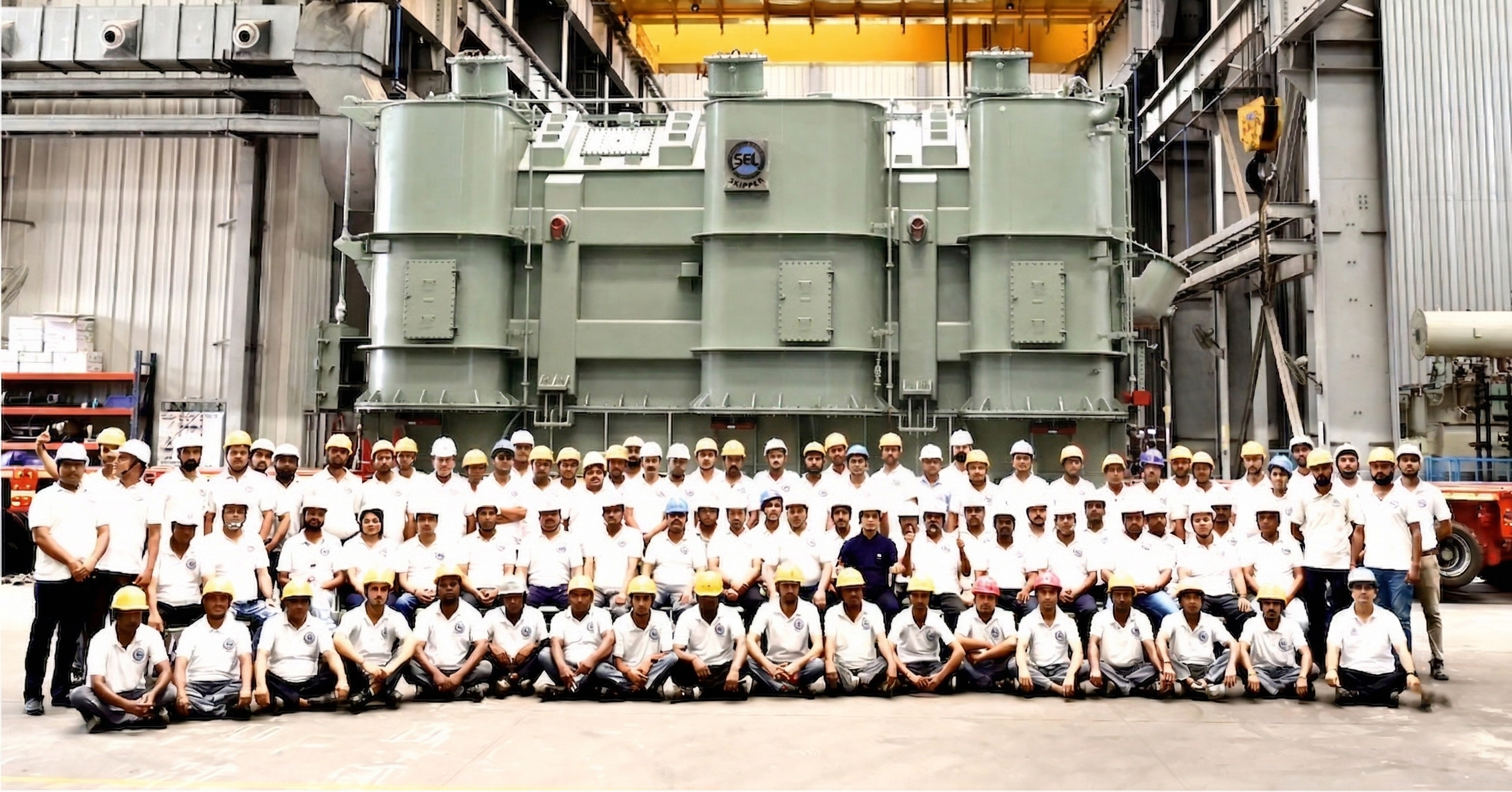 Group of people posing in front of a large industrial transformer with a yellow crane labeled 'REVA' in an industrial setting.
